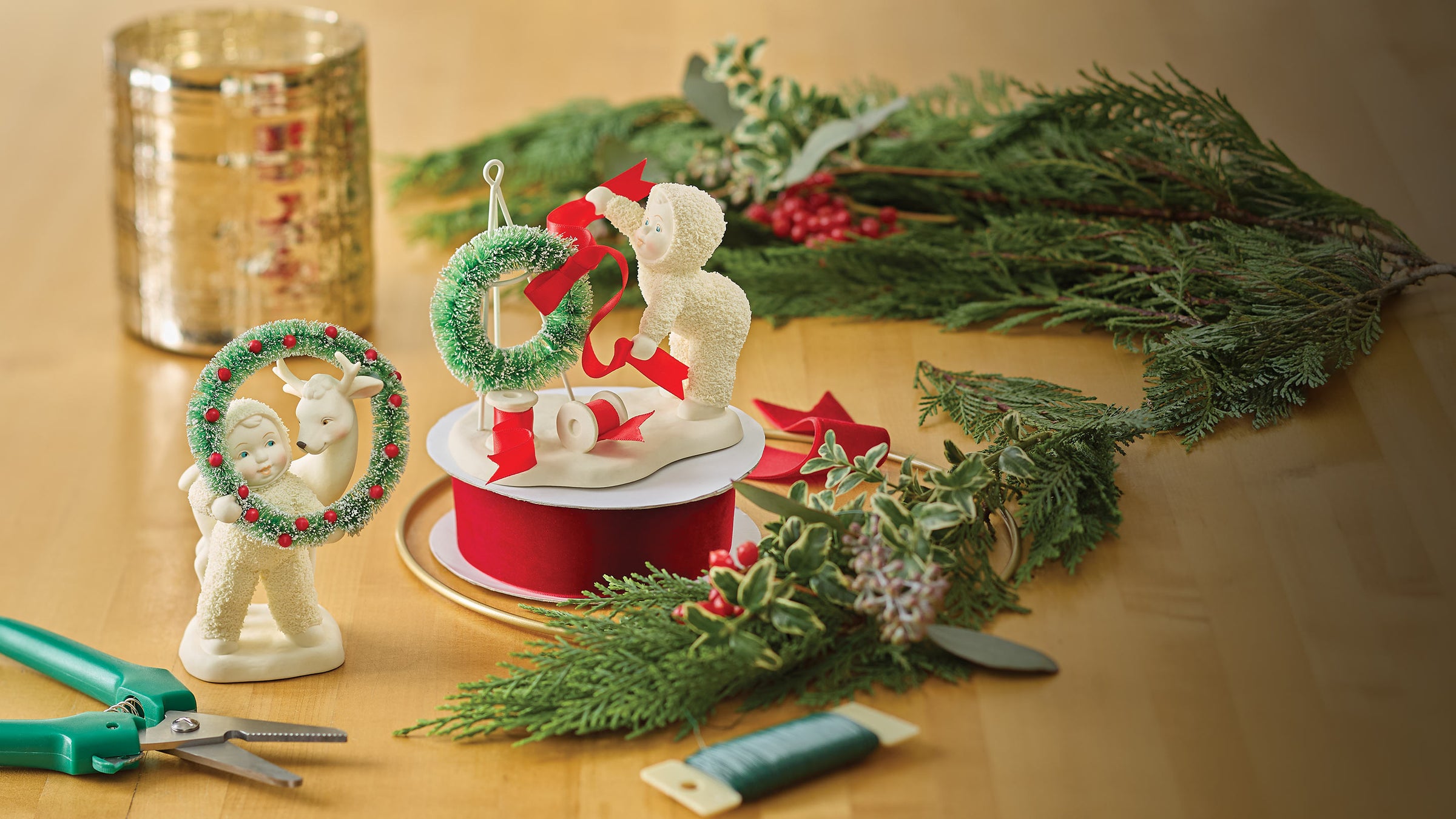 Decorative figurines with wreaths and ribbons on a table with greenery and a candle.