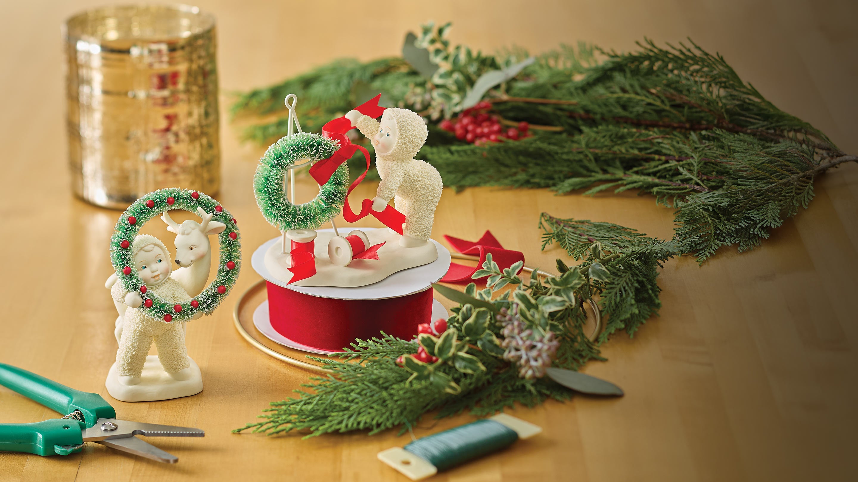 Decorative figurines with wreaths and ribbons on a table with greenery and a candle.