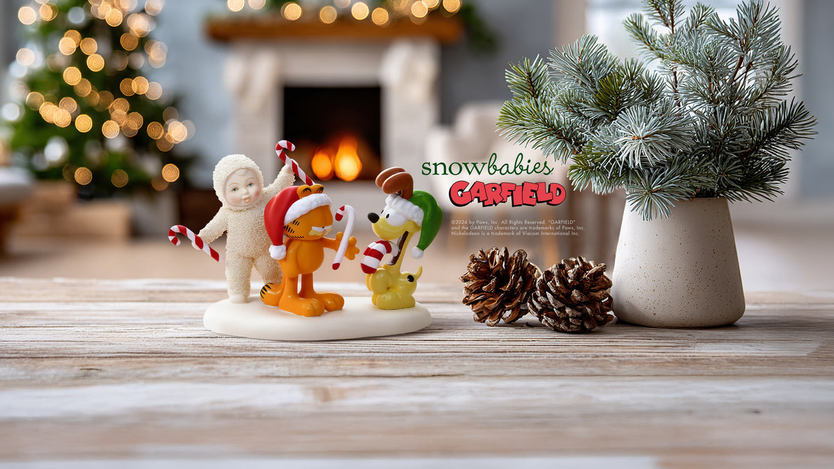 A set of four Snowbabies Garfield holiday figurines displayed on a wooden tabletop with a cozy fireplace, Christmas tree, and potted pine branch in the background. The figurines include Garfield wearing a Santa hat, his cat-like companion in a snowsuit, and other festive characters.