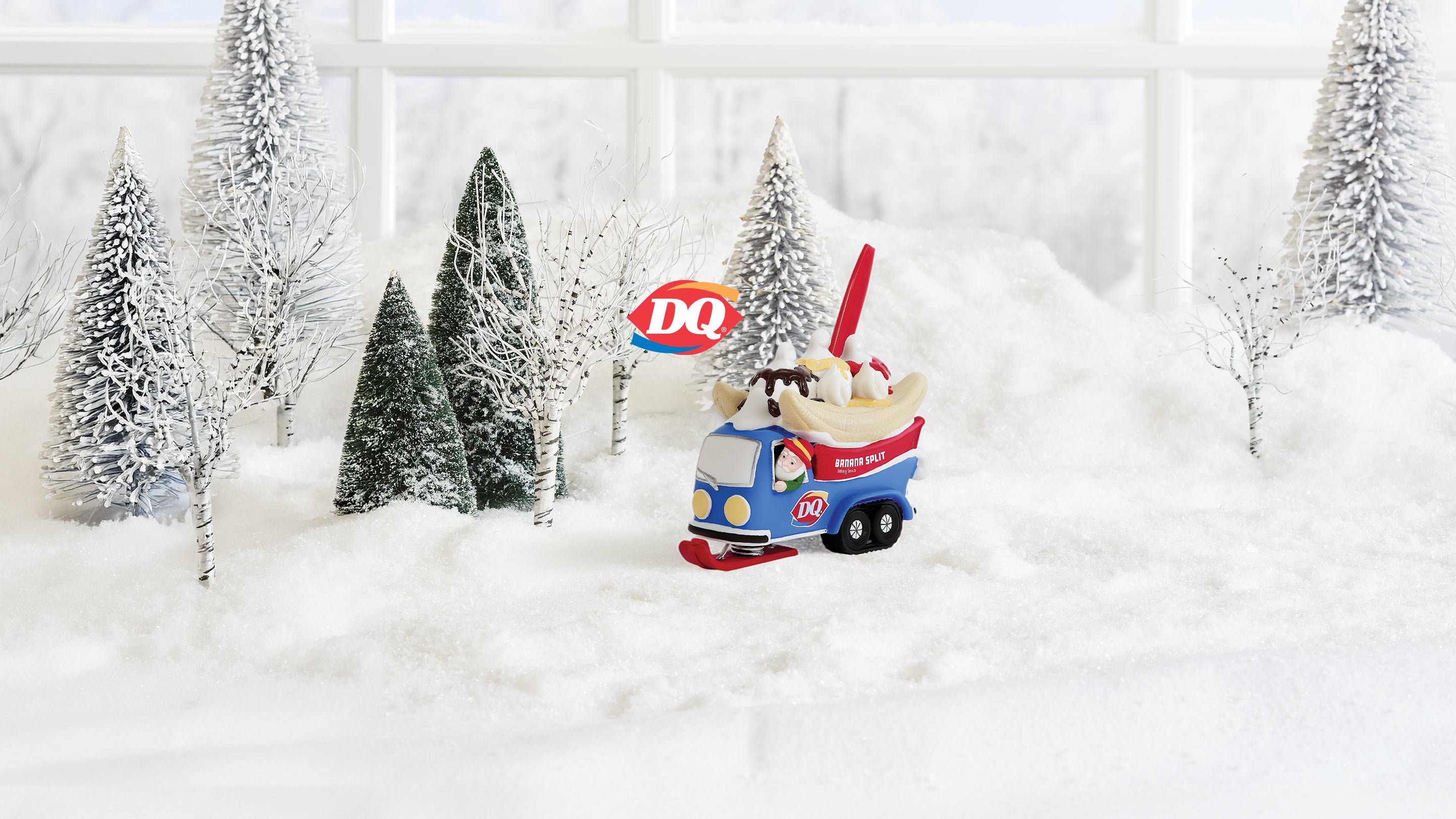 A small Dairy Queen toy truck figurine sitting in a snowy miniature scene with small decorated trees and a blurred window background.