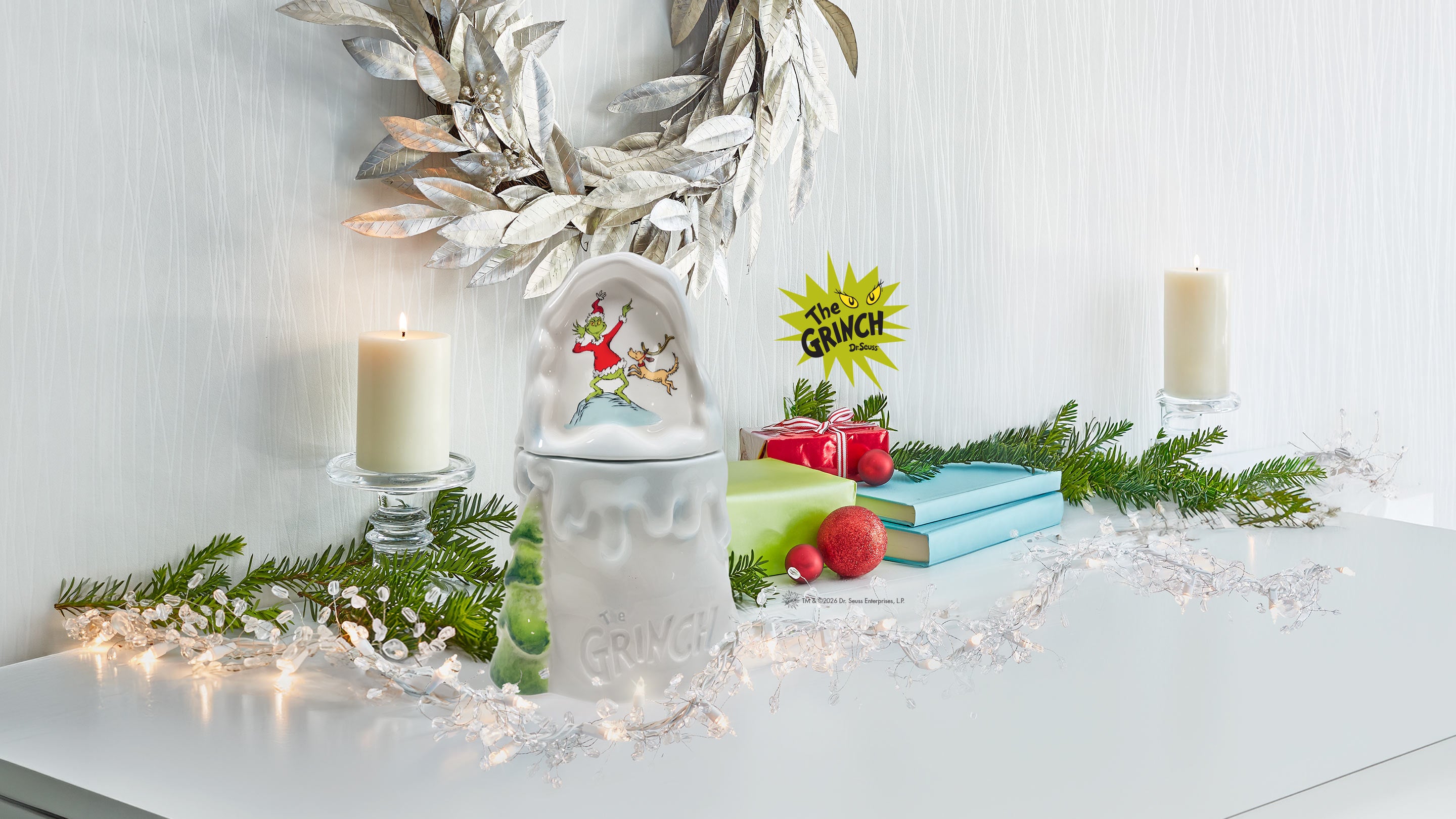 A white ceramic Christmas ornament holder with a Santa Claus and reindeer illustration, styled on a white table with candles, evergreen branches, fairy lights, gift boxes, and a Grinch sign against a white wall.