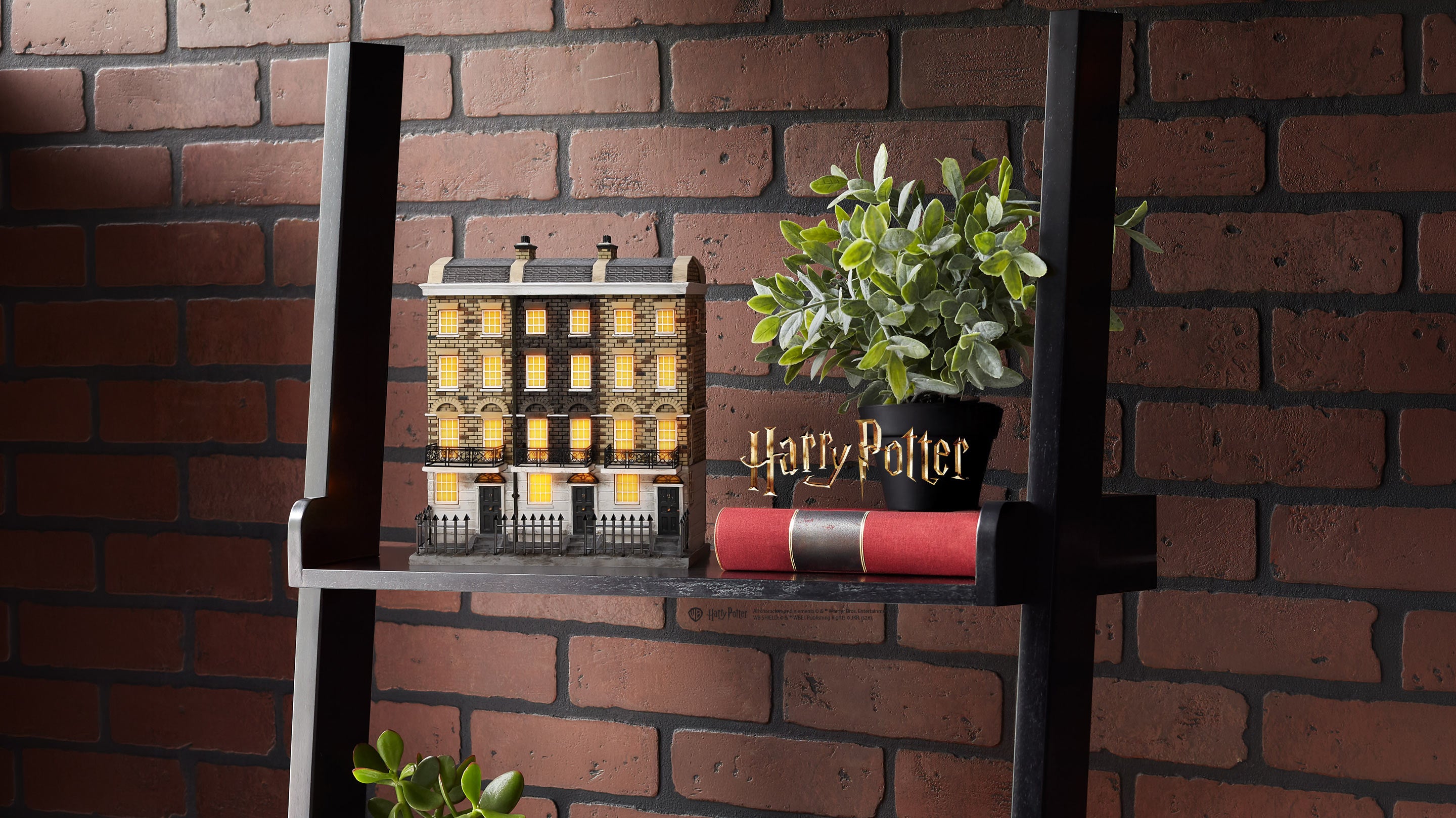 A decorative miniature model of Diagon Alley from Harry Potter, styled on a black metal shelf against a red brick wall background, next to a potted green plant and a red and black Harry Potter book.