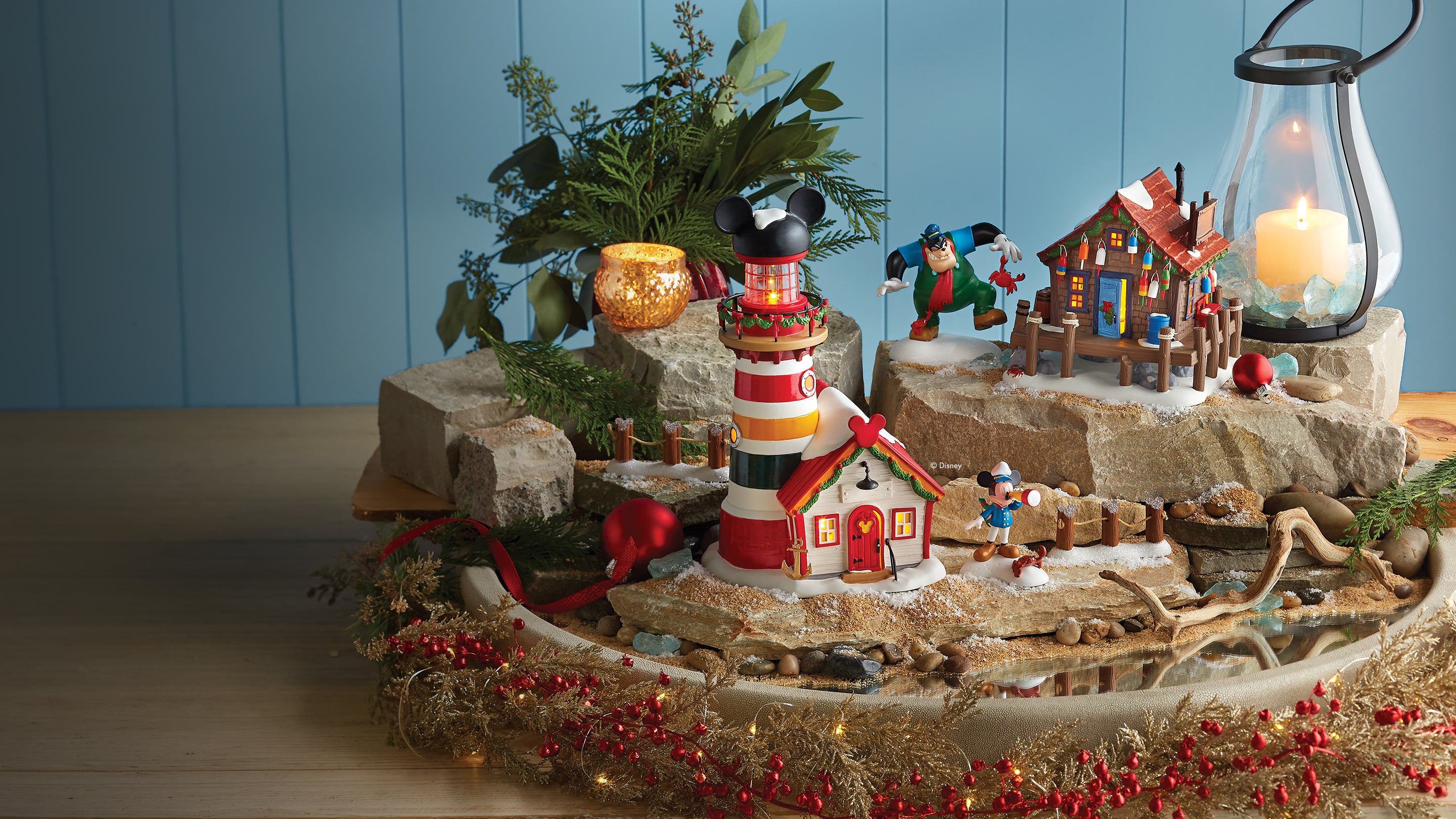 A decorative tabletop holiday display featuring a red-and-white lighthouse with Mickey Mouse ears, a small cottage, a pirate figure, rocks, evergreen branches, red berries, and a lit lantern against a blue paneled background.