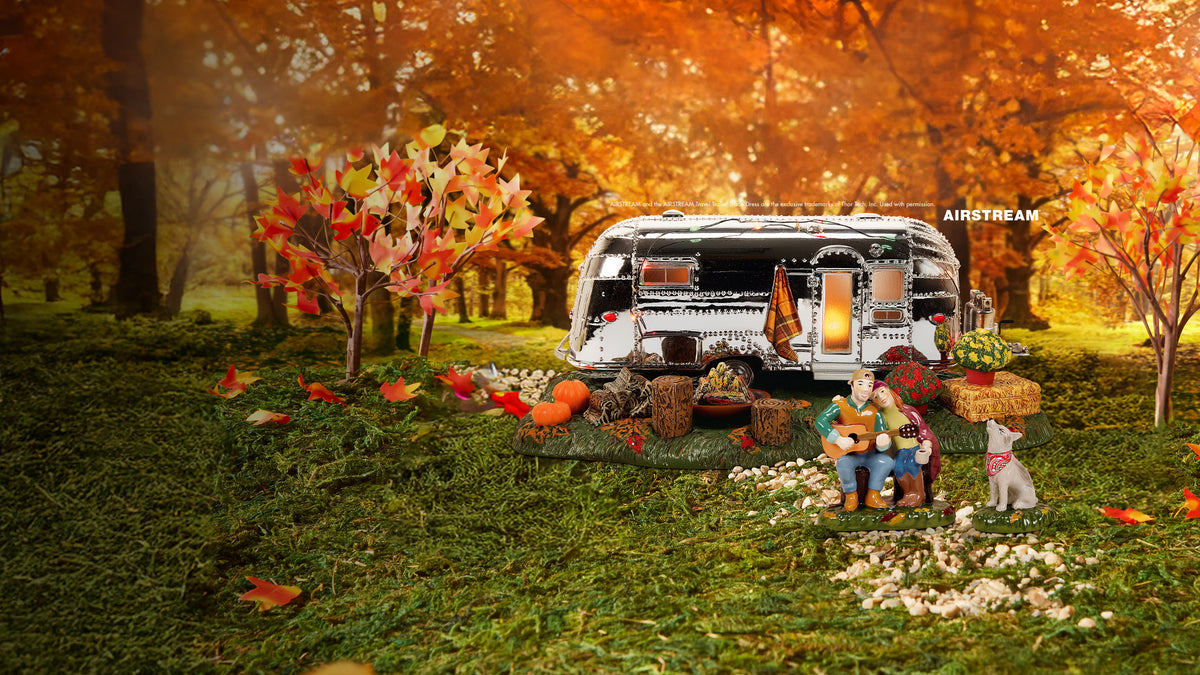 A miniature camping trailer setup in an autumn forest scene, with a small figurine of a couple playing guitar and singing, a dog, baskets of fruit, and warm orange foliage in the background.