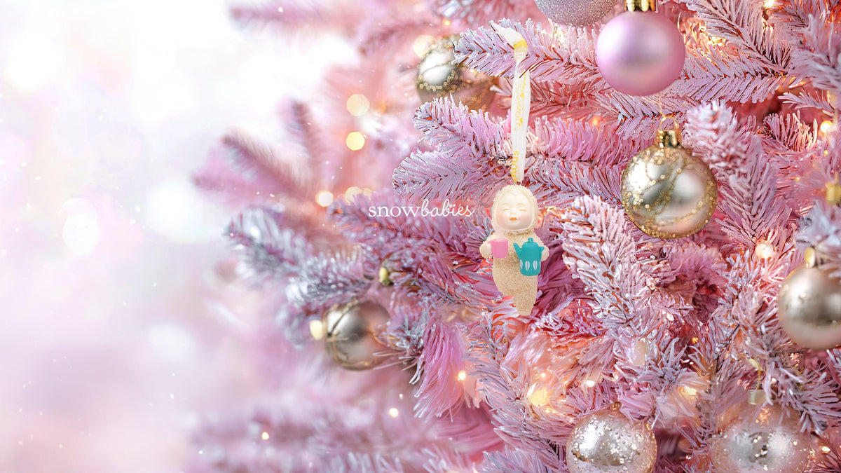 A close-up of a pink Christmas tree decorated with pastel pink and silver ornaments, warm white string lights, and a small hanging angel ornament. The background is a soft, out-of-focus pink bokeh.
