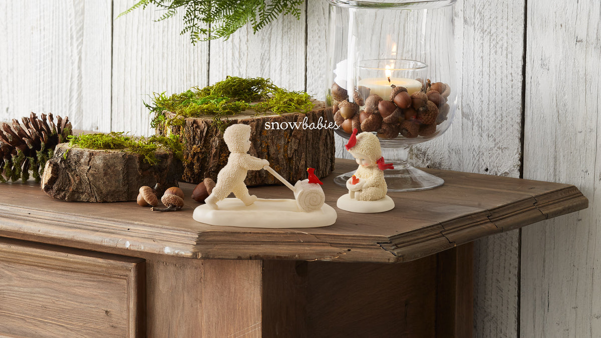A styled holiday tabletop display featuring cream-colored snowman figurines, one pushing a small sleigh and one sitting, alongside mossy logs, pinecones, and a glass jar candle filled with acorns against a white wood plank background.