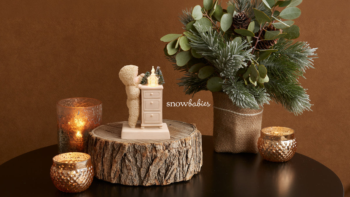 A decorative holiday figurine of a couple embracing in front of a small cabinet, displayed on a rustic wood slice pedestal. The scene is styled with a burlap-wrapped greenery arrangement, gold-toned candle holders, and a warm brown background.
