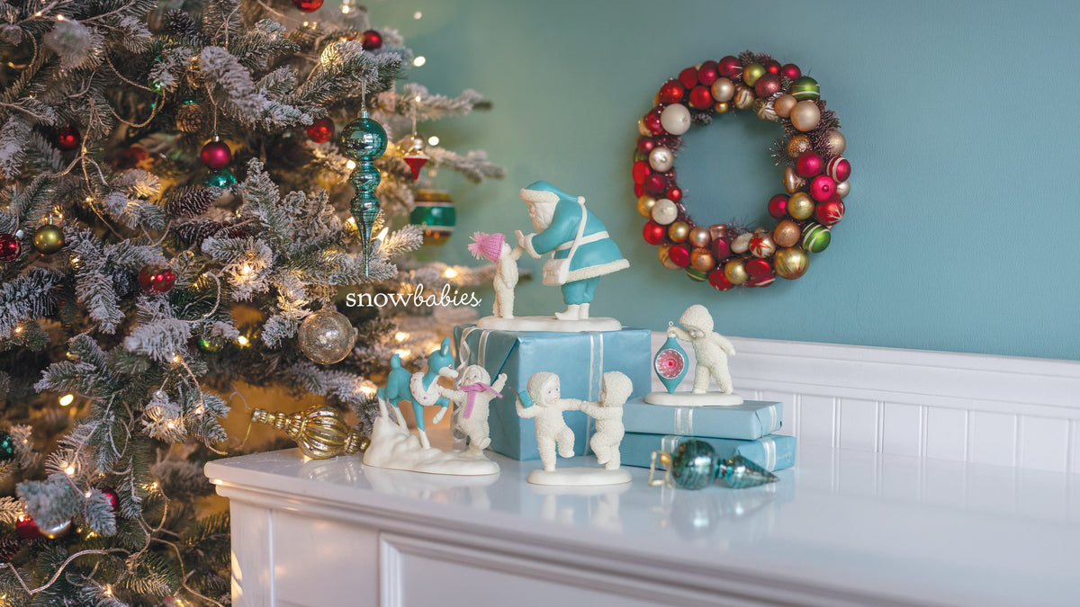 A collection of vintage-style ceramic Christmas figurines displayed on a white mantelpiece next to a decorated flocked Christmas tree. The set includes white angel and baby figures, a blue-clad Santa figure, and a small clock figurine, arranged with light blue gift boxes.