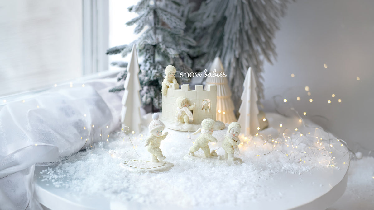 A set of off-white miniature winter-themed figurines, including small snowmen, a castle with a figure on top, and cone-shaped trees, arranged on a white surface with faux snow, fairy lights, and a blurred Christmas tree in the background.