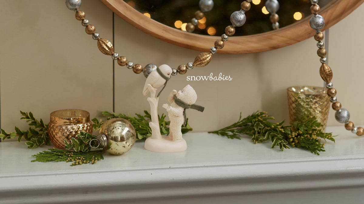 A holiday-themed tabletop display featuring a cream-colored snowman figurine holding a small snowball, flanked by green pine garlands, gold and silver ornaments, and a beaded garland draped over a mirror. The text 'snowbabies' is visible in the center.