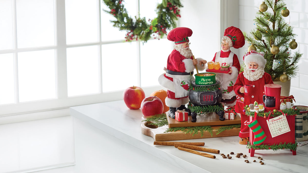 A festive Christmas tabletop display with three Santa Claus figurines, a small tree, ornaments, apples, cinnamon sticks, and gift boxes on a white windowsill.
