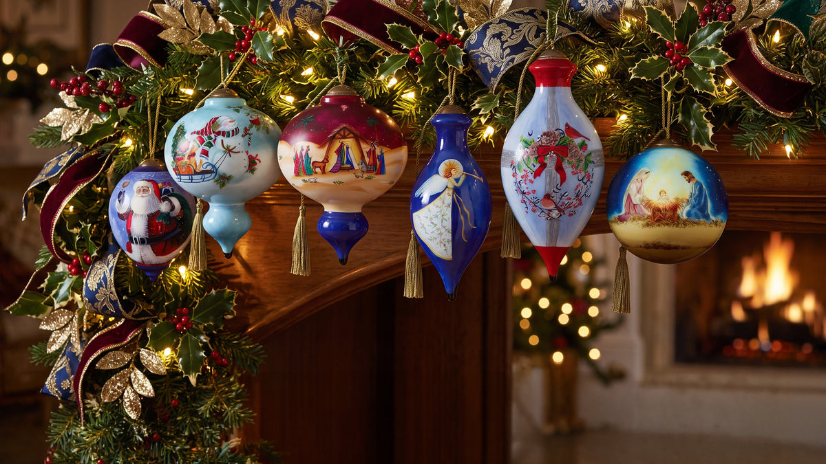 A set of hand-painted Christmas ornaments hanging from a decorated Christmas tree branch in front of a warm fireplace. The ornaments feature various festive designs including Santa Claus, a nativity scene, an angel, and a bride and groom, in colors of blue, red, white, and gold.