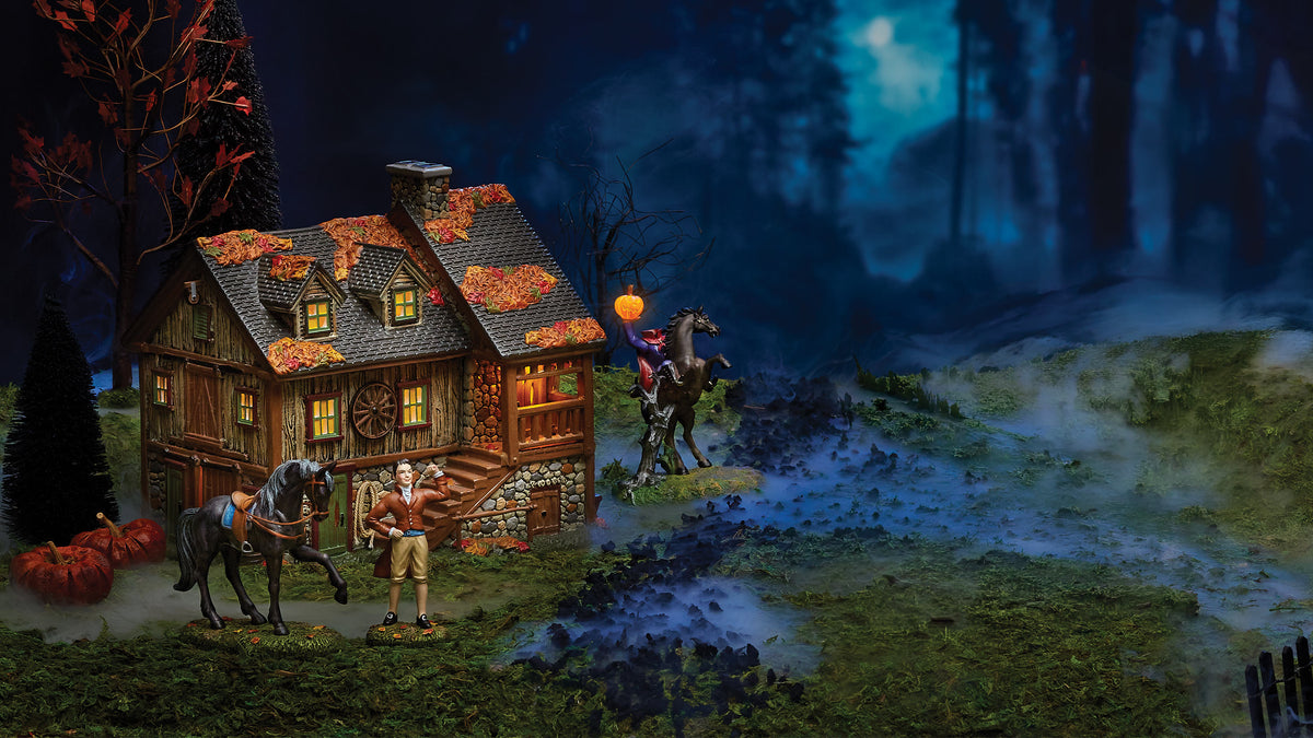 A miniature Halloween village scene featuring a small lit cottage with autumn leaves on the roof, a standing figurine, a horse figurine, and a rider on horseback, set against a misty blue night forest backdrop.