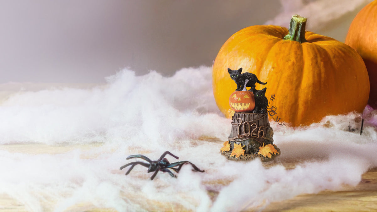 Halloween-themed tabletop scene with orange pumpkins, white faux cobwebs, a black spider toy, and a small decorative bell figurine on a wooden surface.