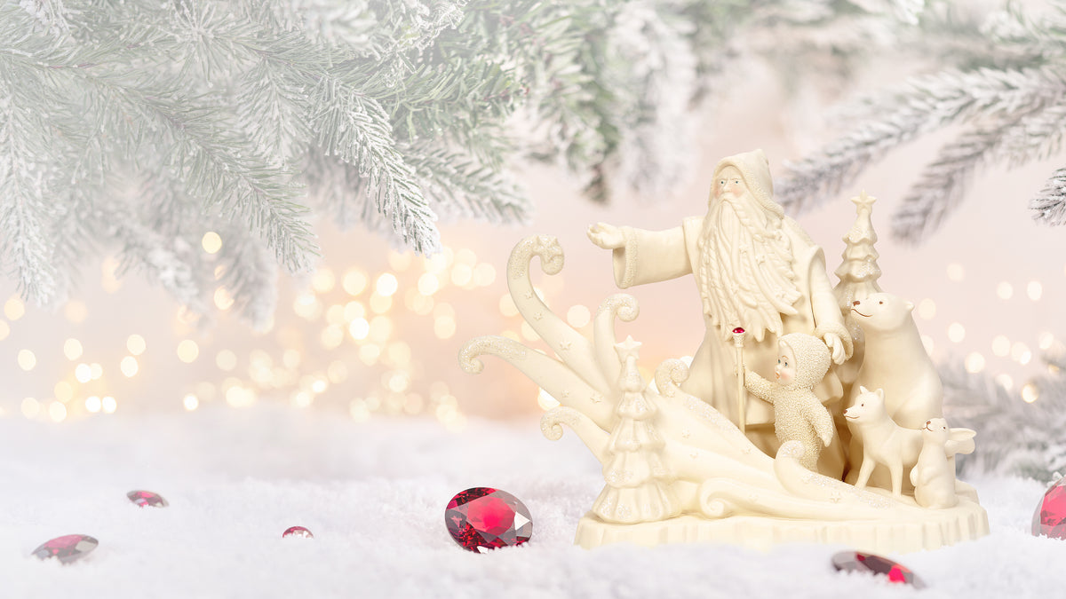 A small cream-colored Christmas nativity figurine set with a Santa figure, baby Jesus, Mary, Joseph, and animals, displayed on a snowy surface with red decorative gems and blurred pine branches in the background.